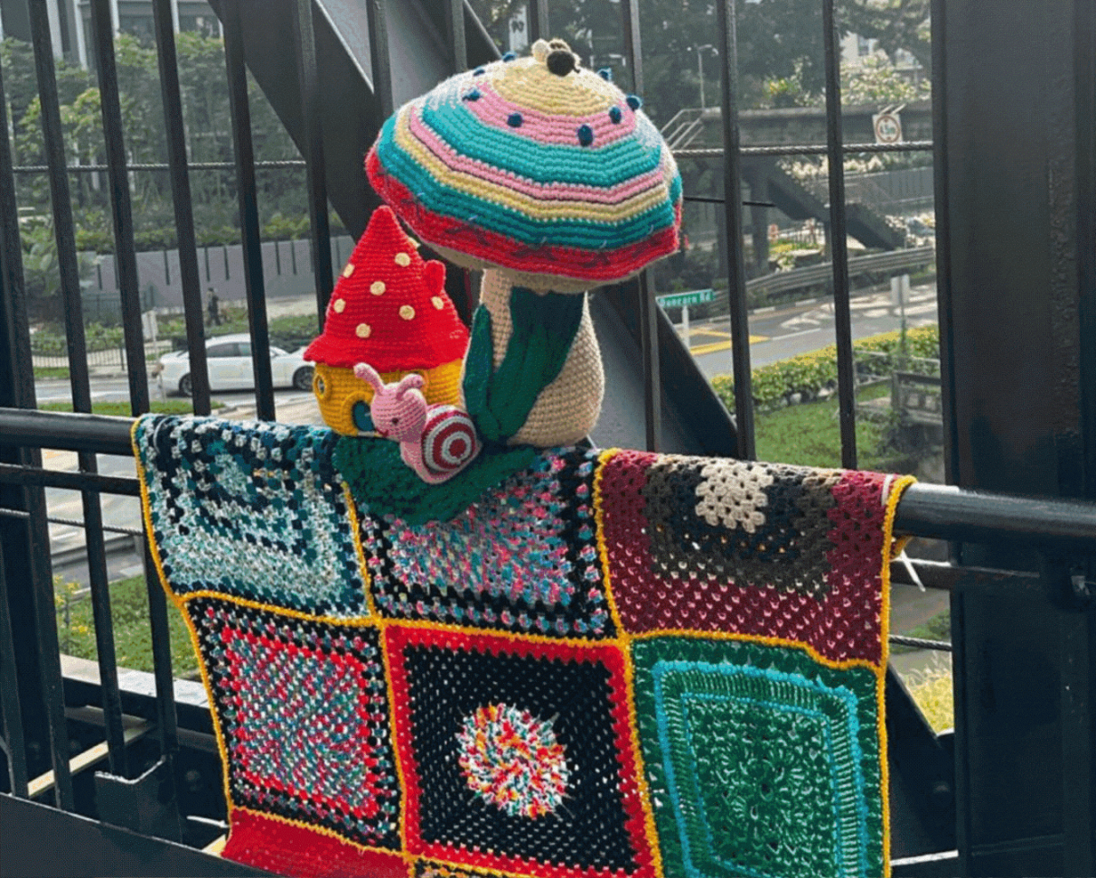 Colourful crochet installations on a railing: a patchwork blanket, a large mushroom, a red toadstool house, and a small snail figurine